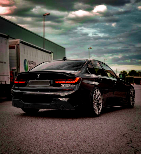A black car with illuminated rear lights is parked on an empty street. Large wheels and a sleek design stand out. Nearby are cargo trailers under a cloudy sky.