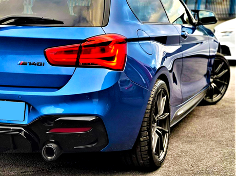 A blue sports car labeled "M140i" is parked, displaying dual exhausts and vibrant taillights, set on a paved surface with reflections of the sky and surroundings.