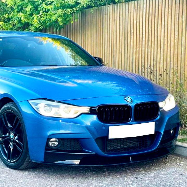 A blue car is parked with headlights on beside a wooden fence, surrounded by greenery.