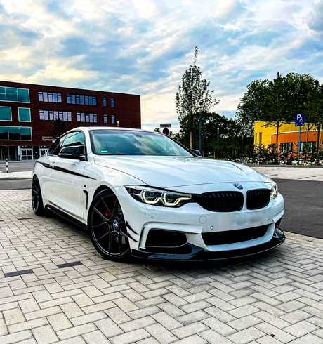 A white BMW car parked on a paved area in front of modern buildings under a cloudy sky. Trees and a sign with a "P" symbol are visible in the background.