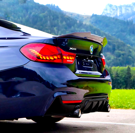 A sleek blue BMW car parked, highlighting its tail lights and exhaust system, with a forested mountain landscape in the background. Text: "XDrive 440i."
