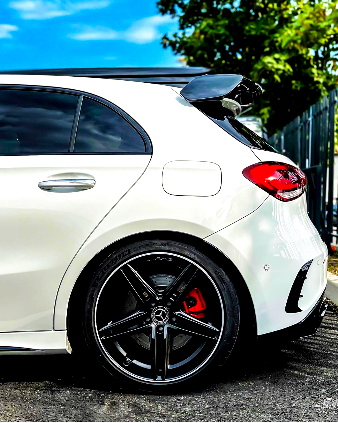 A white car is parked, showcasing its rear side with sleek black alloy wheels and a roof spoiler, amid a sunny day with a blue sky and some greenery nearby.
