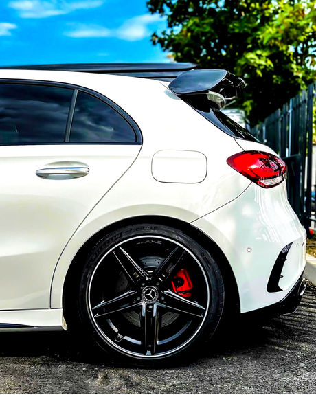 A white car is parked, showcasing its rear side with sleek black alloy wheels and a roof spoiler, amid a sunny day with a blue sky and some greenery nearby.