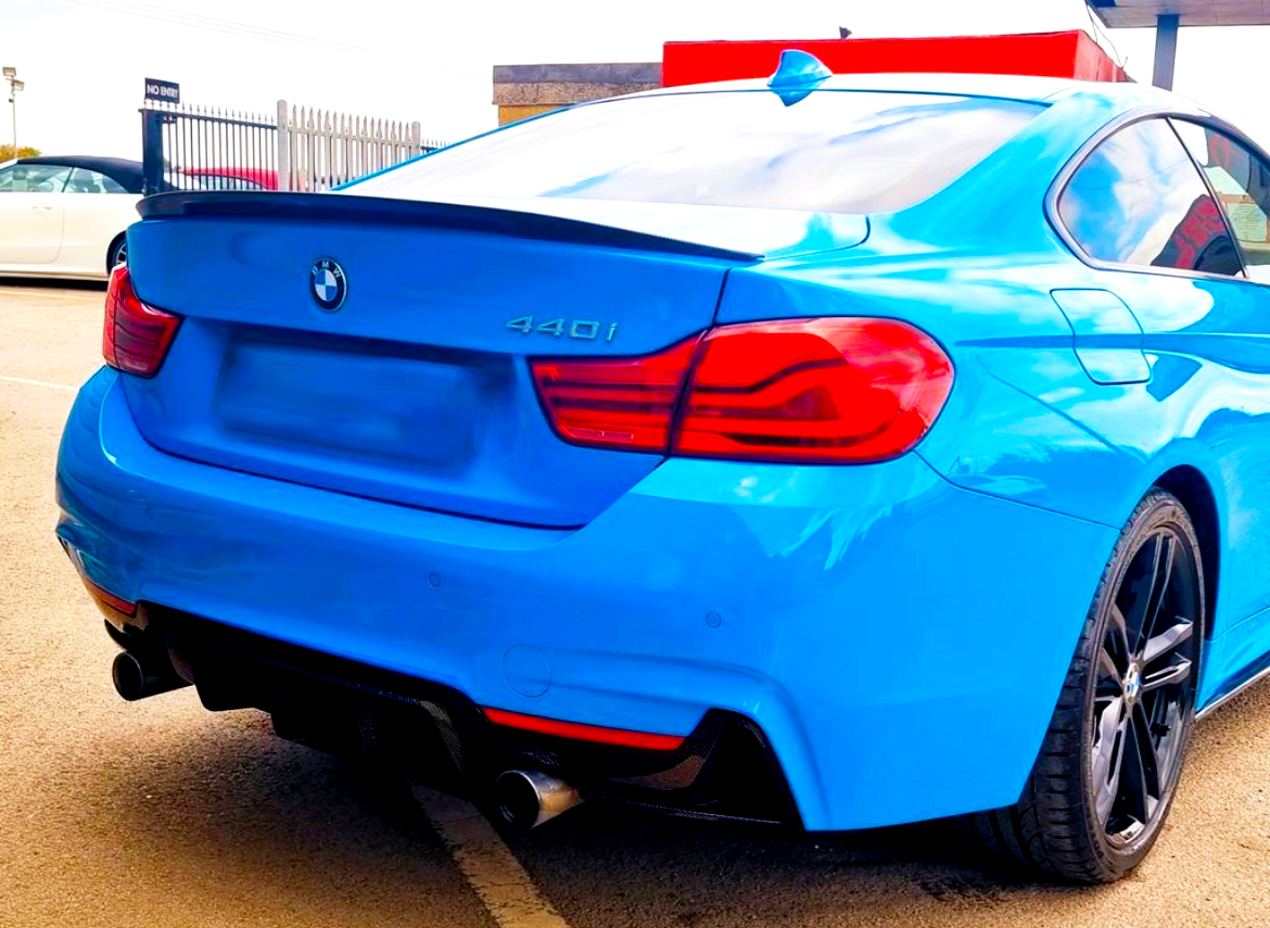 A blue BMW 440i car is parked on a light-colored surface, featuring dual exhausts and red-taillights. A fenced area and another parked car are visible in the background.