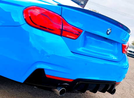A blue BMW 440i with dual exhaust is parked on asphalt. Its distinctive rear lights and emblem are visible in bright daylight. Another car is partially visible nearby.
