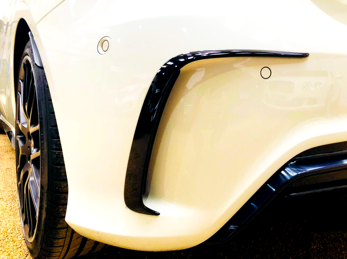 A car's white rear bumper with parking sensors and a black accent curves around the wheel. The scene is set inside a well-lit vehicle display area.