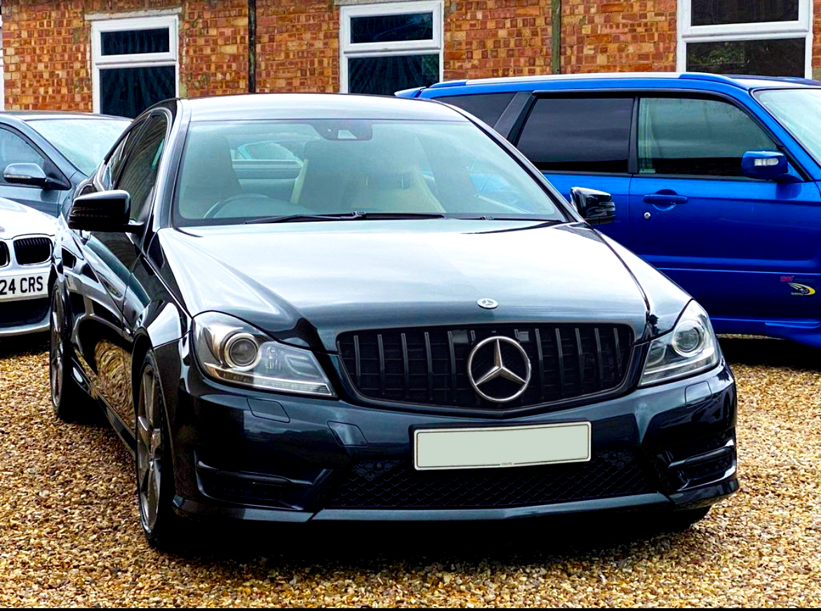 A black Mercedes-Benz is parked on gravel. It is flanked by a silver car on the left and a blue car on the right. The background features a brick building with windows.