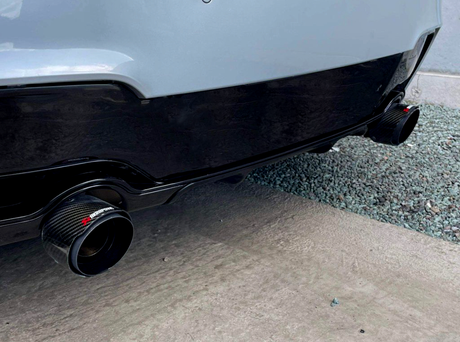 Exhaust pipes with carbon fiber tips are affixed to the rear of a car, positioned above gravel and pavement. The text "Akrapovič" is visible on the left pipe.