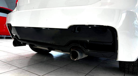 A white car's rear bumper with a black diffuser features dual exhaust pipes, located indoors, with another red vehicle partially visible in the background.