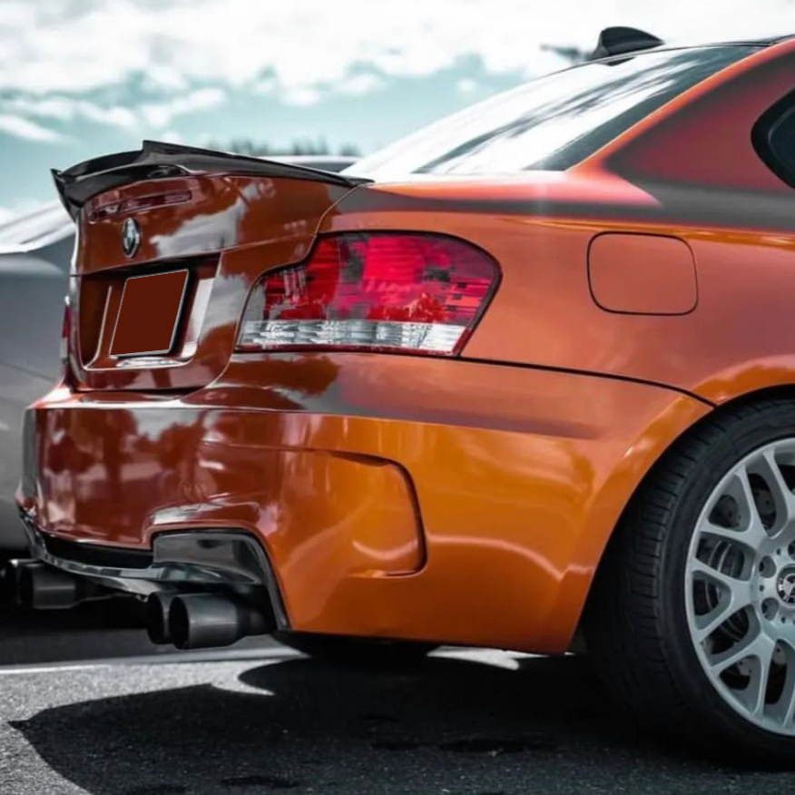 An orange BMW car is parked, showcasing its rear left side with tail lights, spoiler, and dual exhaust against a cloudy sky backdrop.