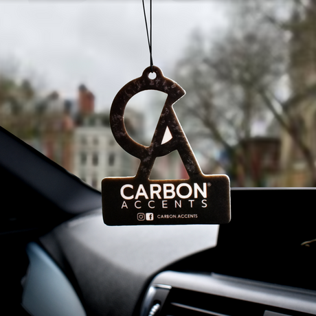 A car air freshener with a stylized "CA" logo hangs on a string in front of the windshield. Text reads: "CARBON ACCENTS" with social media handle @CARBON.ACCENTS. Background shows blurred buildings.