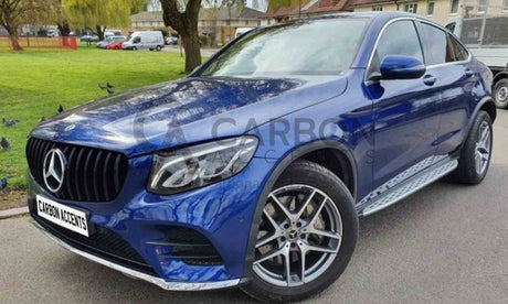 A blue Mercedes car parked on a road with a side view of large alloy wheels. Background features a grassy area with trees and buildings. Text: "CARBON ACCENTS" on the license plate.