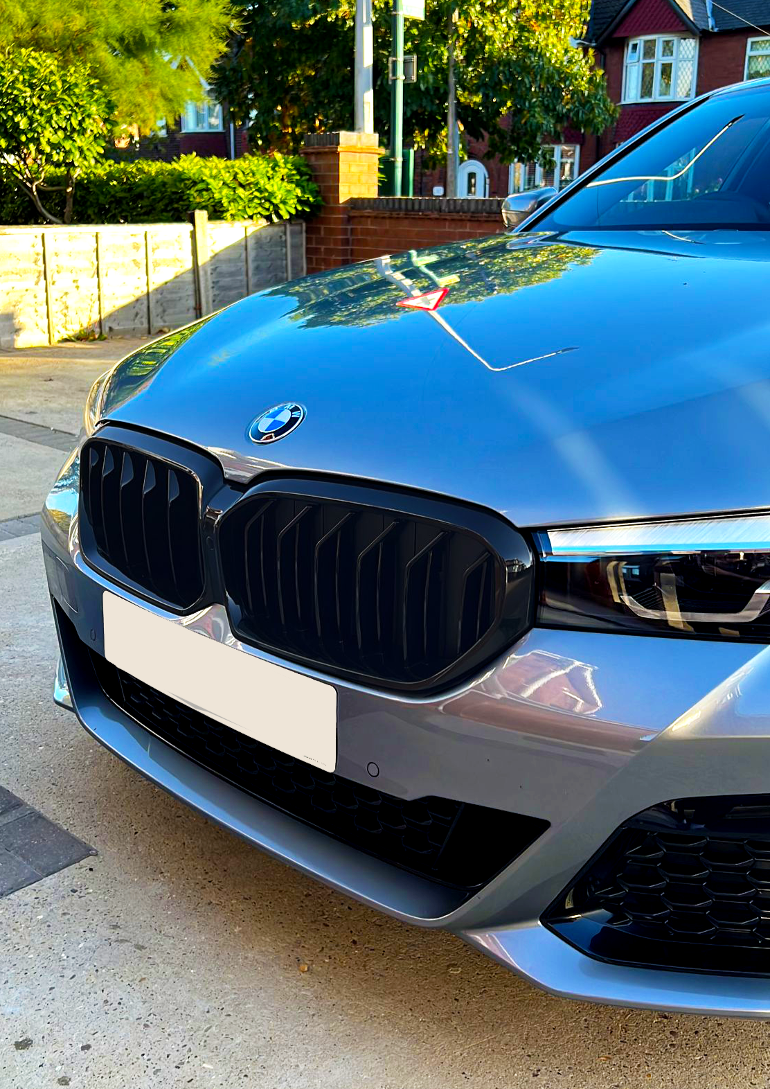 A silver BMW car is parked on a driveway, with a prominently visible grille and headlight. Trees and brick houses form the background under a clear blue sky.