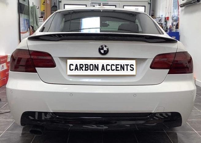 A white BMW is parked in a garage. It features a "CARBON ACCENTS" license plate. The setting includes tools and equipment on shelves.