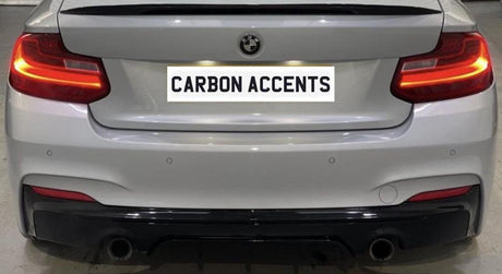 A white BMW car's rear, illuminated taillights, in a garage setting. License plate reads "CARBON ACCENTS." The car has a black lower bumper and dual exhaust pipes.