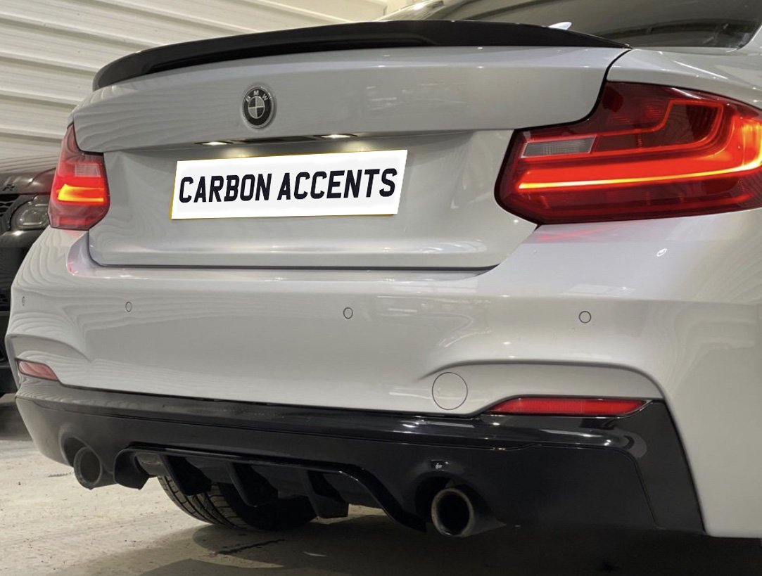 A white car with illuminated taillights, featuring dual exhausts and a sleek carbon diffuser, is parked indoors. License plate text reads: "CARBON ACCENTS."