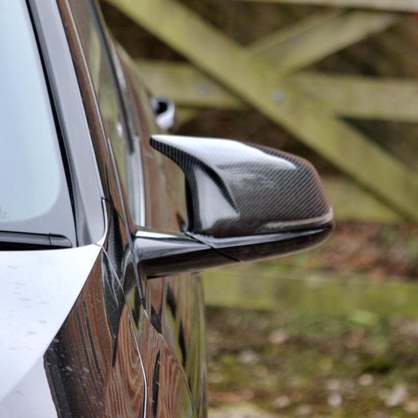 Car side mirror with carbon fiber casing reflecting the car's sleek body, set against a blurred wooden fence background.