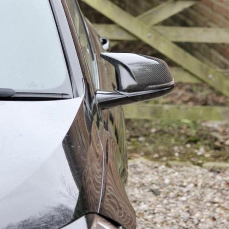 A car's glossy black side mirror is angled outward, reflecting the adjacent environment. The background reveals a gravel driveway and a wooden fence in an outdoor setting.