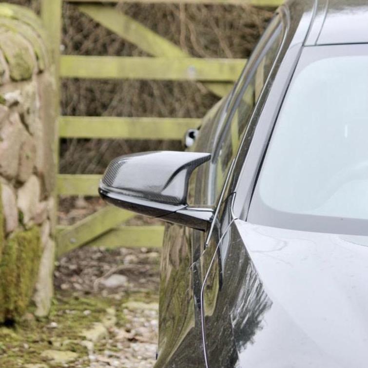 A black car is parked on a gravel driveway next to a stone wall and a wooden gate with a natural backdrop of trees.
