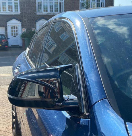 A blue car's side mirror, reflecting nearby houses, is parked on a residential street with brick houses and white doors under a clear sky.