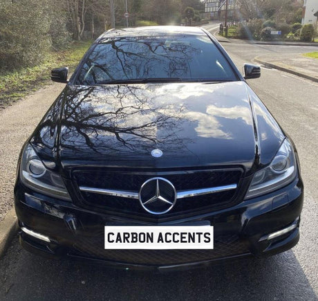 A black Mercedes car is parked on a street, reflecting surrounding trees on its shiny hood. The license plate reads "CARBON ACCENTS." The street has greenery on the side.