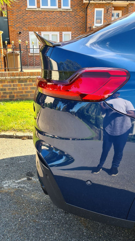 A blue car's rear with a red taillight is parked on a street. A person's reflection is visible on the car. A brick house and grass are in the background.