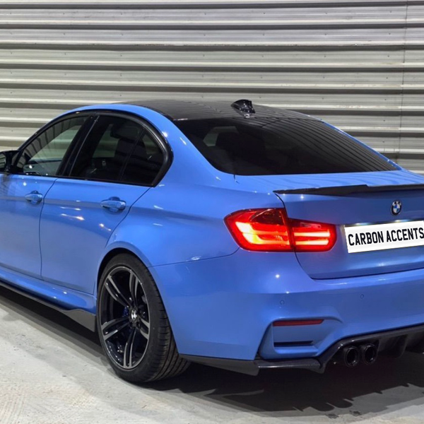 A blue BMW sedan is parked indoors, showcasing its sporty rear with illuminated taillights and carbon accents. The surroundings include a metal-paneled wall. The license plate reads "CARBON ACCENTS."