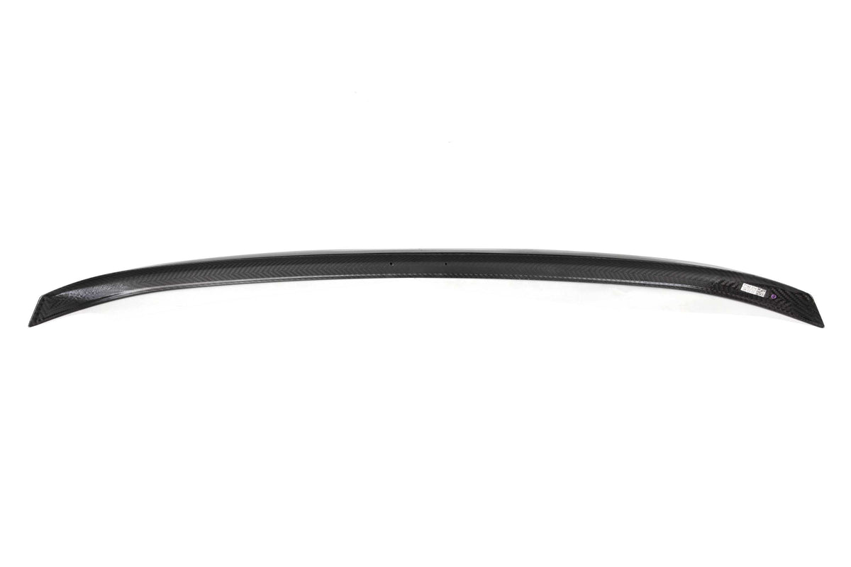 A curved, black carbon fiber car spoiler rests on a white background, displaying a smooth, textured pattern and a small label on the right side.