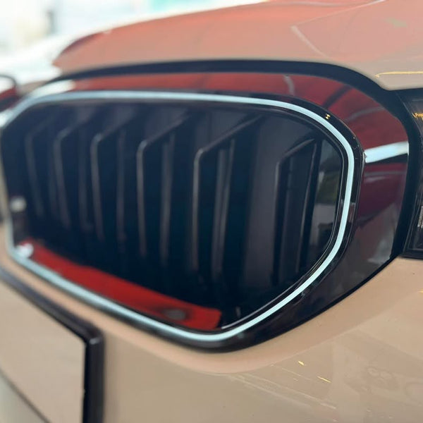 Car grille with vertical slats, bordered by chrome, situated on a beige vehicle. Right headlight partially visible, in a bright showroom environment.