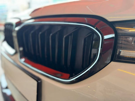 Car grille with vertical slats, bordered by chrome, situated on a beige vehicle. Right headlight partially visible, in a bright showroom environment.