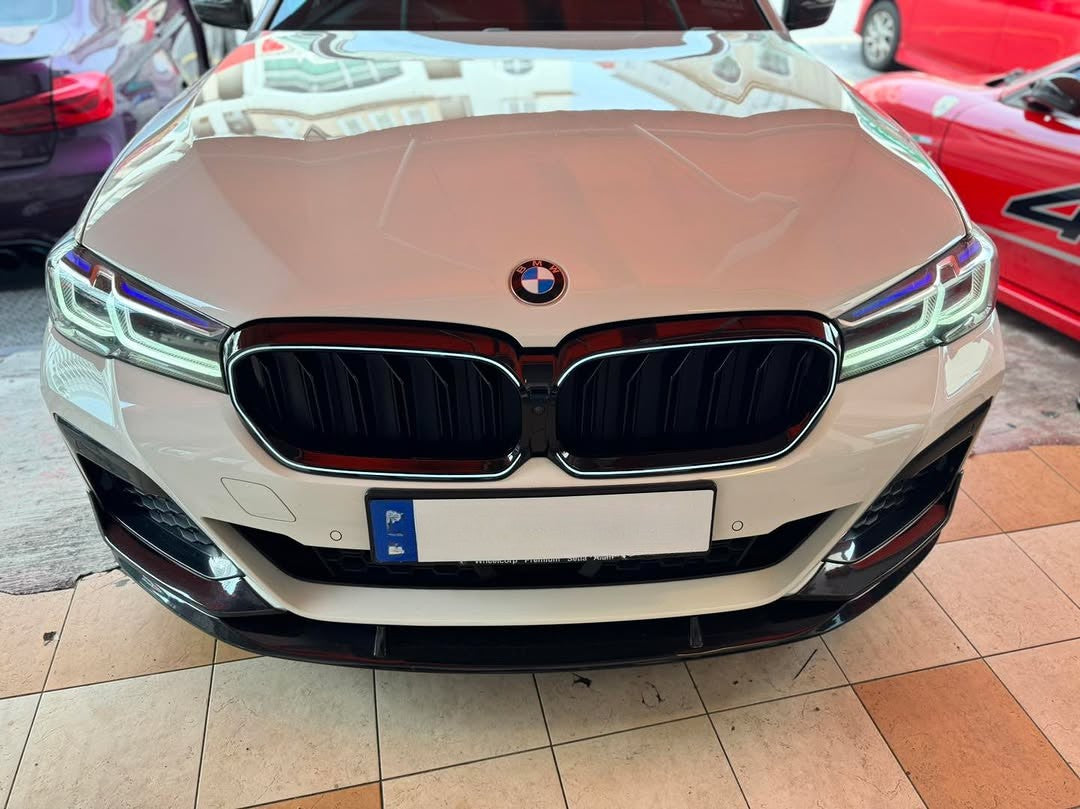 A white BMW car front is parked on a tiled floor, with reflections of buildings on the hood. Nearby are partially visible red and purple cars.