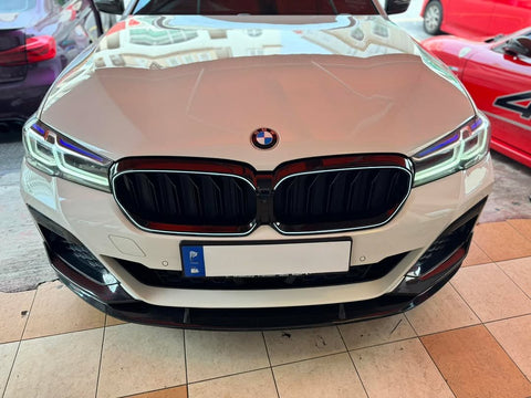 A white BMW car front is parked on a tiled floor, with reflections of buildings on the hood. Nearby are partially visible red and purple cars.