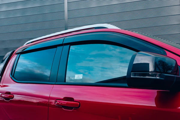 A red vehicle is parked with closed windows reflecting a cloudy sky. The background features a corrugated metal wall. A side mirror protrudes, and no text is visible.