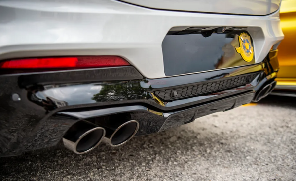 A car's rear, featuring dual exhaust pipes, is parked on asphalt. It has a glossy black diffuser and a blurred badge with a star shape on the left side.