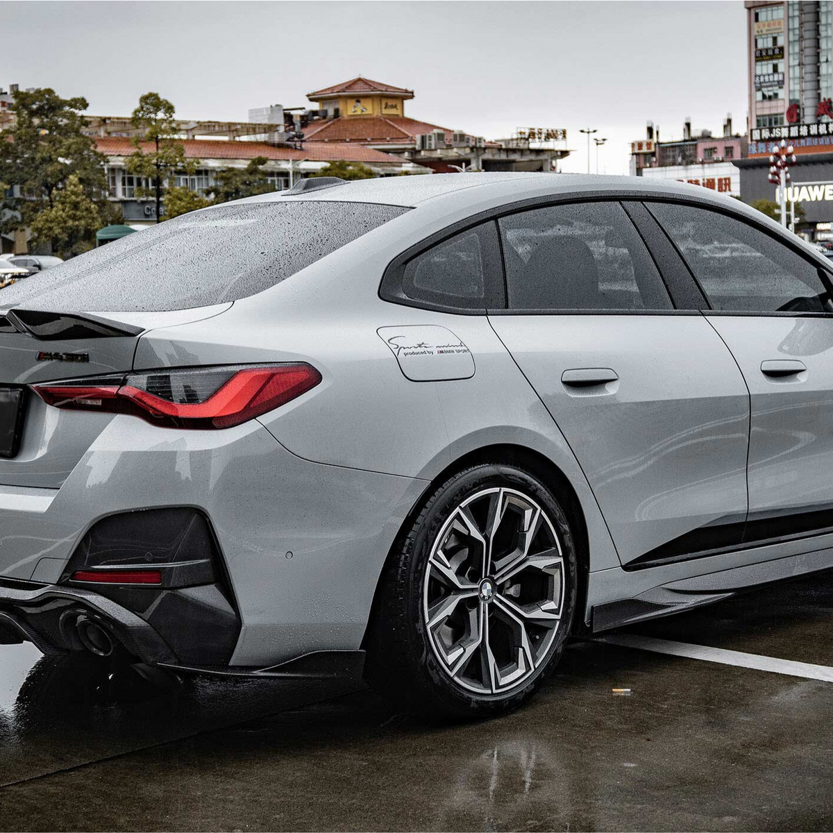 A silver car with BMW logo is parked on a wet pavement in an urban environment, featuring sleek design and sporty wheels. Surrounding buildings display signage, including "Huawei."