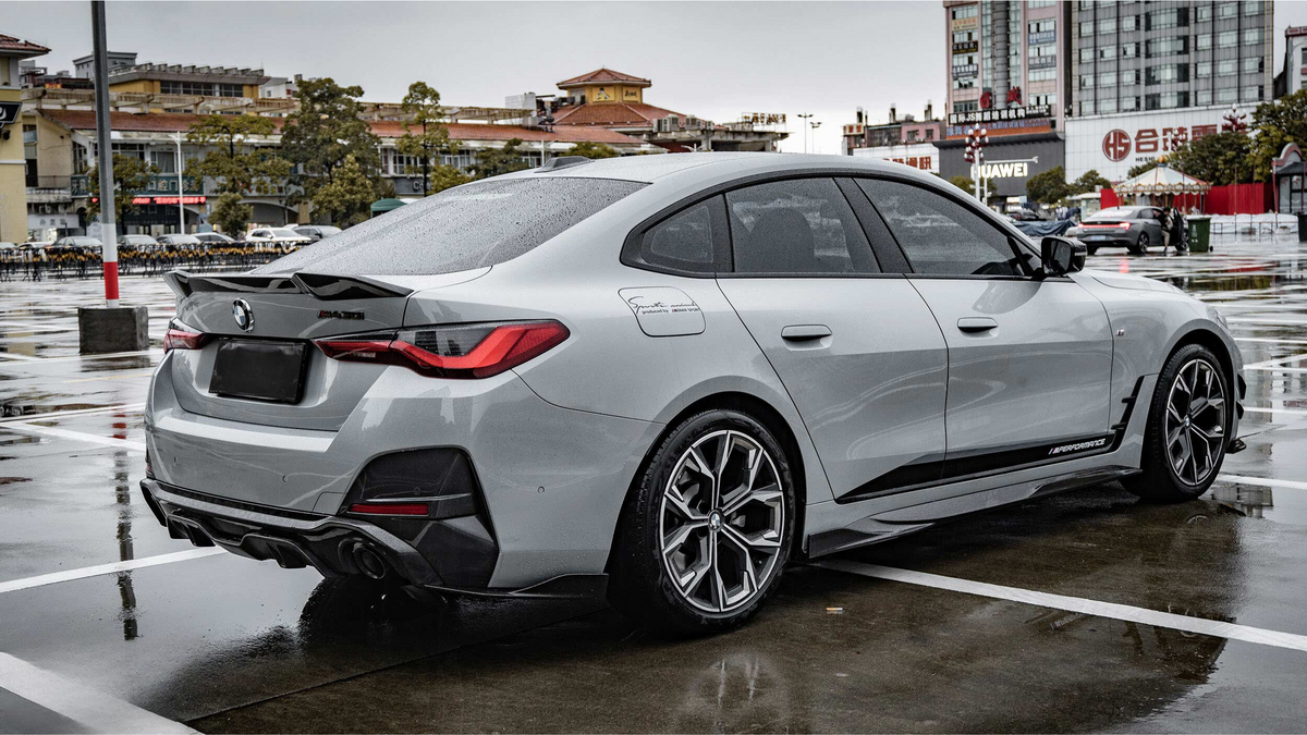 A silver car with BMW logo is parked on a wet pavement in an urban environment, featuring sleek design and sporty wheels. Surrounding buildings display signage, including "Huawei."