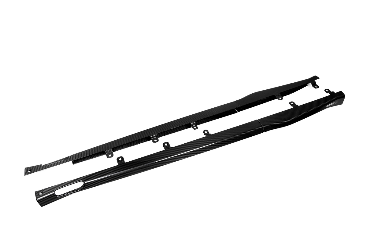 Two long, black carbon fiber side skirts with mounting brackets lie side by side on a plain white background. The word "Amuse" is visible on one end.
