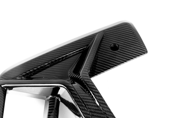 A glossy carbon fiber automotive part featuring a geometric design with sharp angles, showcased against a plain white background. The surface has a distinctive woven pattern and a small circular hole.