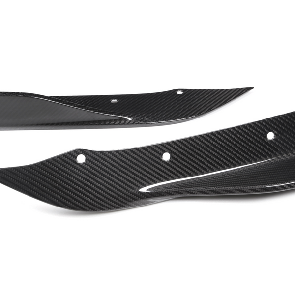 Two carbon fiber car parts, possibly splitters or fins, lie on a white background. They have aerodynamic shapes with several bolt holes for mounting.