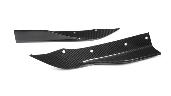 Two carbon fiber car parts, possibly splitters or fins, lie on a white background. They have aerodynamic shapes with several bolt holes for mounting.