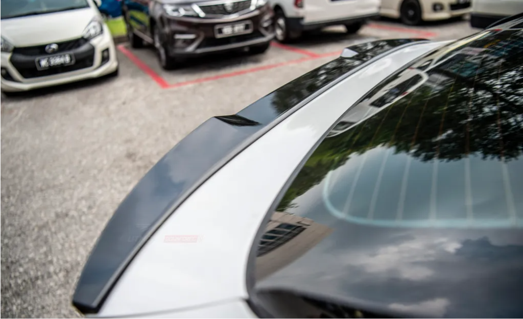 A glossy car, parked, reflects trees and sky on its rear window. Other vehicles are parked nearby in an outdoor lot.