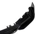 A glossy carbon fiber car accessory, likely a bumper, lies on a plain white background. The item features aerodynamic curves and multiple mounting points for attachment.