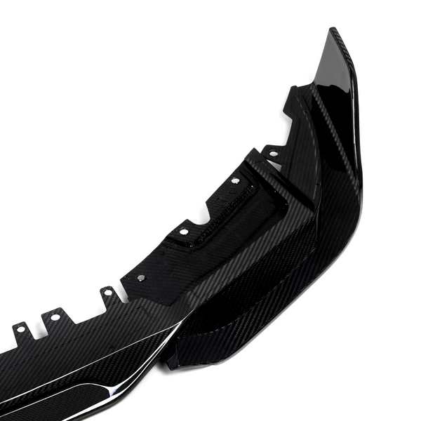 A glossy carbon fiber car accessory, likely a bumper, lies on a plain white background. The item features aerodynamic curves and multiple mounting points for attachment.
