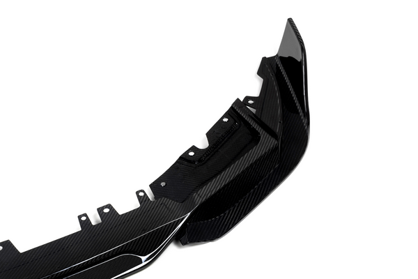 A glossy carbon fiber car accessory, likely a bumper, lies on a plain white background. The item features aerodynamic curves and multiple mounting points for attachment.