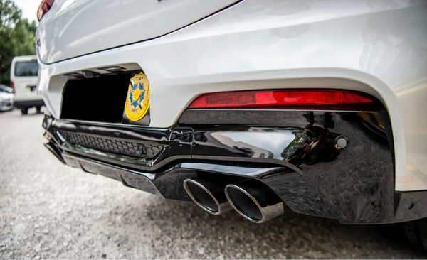Car rear shows black triple exhaust pipes and red taillight, parked on asphalt. Yellow badge reads "CMP DATO" with an emblem. Trees and another vehicle are nearby.