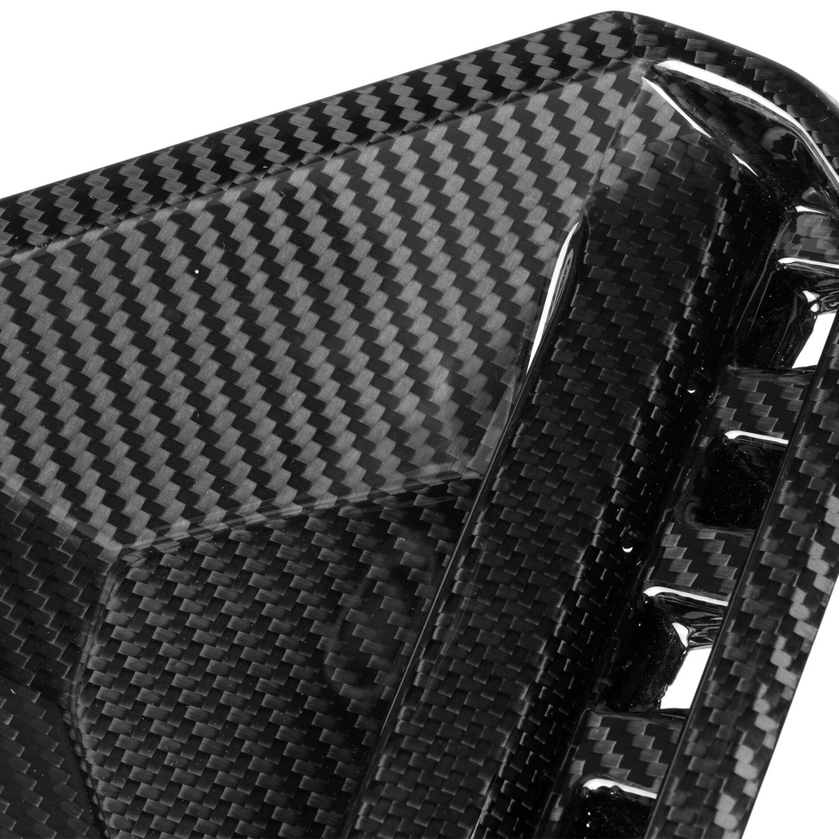 A close-up of a glossy carbon fiber object, likely a car part, with a woven pattern and angular design, featuring small vents or openings along the edge.