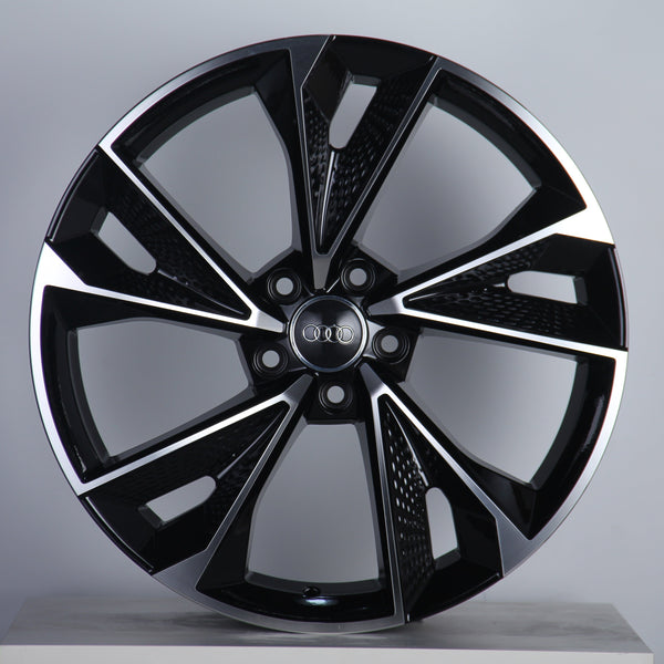 A black and silver alloy wheel with a geometric design and five dual spokes is displayed upright on a neutral background. The center features a logo with four interlocking rings.