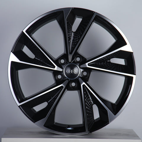 A black and silver alloy wheel with a multi-spoke design is displayed on a neutral background, featuring the Audi logo at its center and intricate detailing on each spoke.