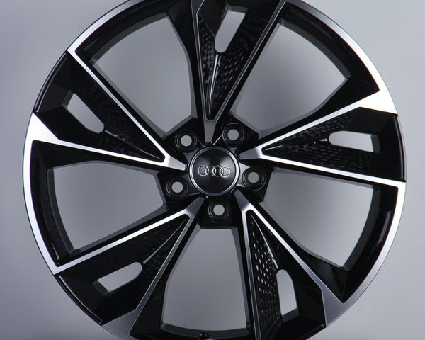 A glossy black car wheel with five split spokes features a central circle displaying four interlinked rings. It rests against a plain gray background, highlighting its modern design.