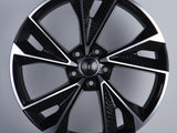 A sleek, glossy black alloy wheel with a multi-spoke design featuring the Audi logo at its center, displayed against a plain gray background on a white platform.